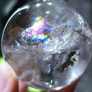 Puede incluir: Una esfera de cristal de cuarzo transparente con inclusiones iridiscentes. La esfera tiene un diámetro de aproximadamente 5 cm y una superficie lisa y pulida.