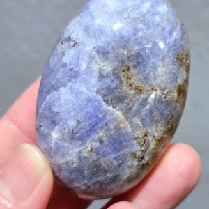May include: A polished, oval-shaped gemstone in shades of lavender and blue, with natural brown and tan inclusions. The stone is held in a hand, showcasing its smooth, tactile surface. This unique stone is a natural mineral specimen.