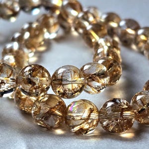 Puede incluir: Dos pulseras de cuentas de color ámbar. Las cuentas redondas y translúcidas presentan inclusiones internas que crean un patrón único. Las pulseras se exhiben sobre una superficie gris claro, mostrando la joyería.