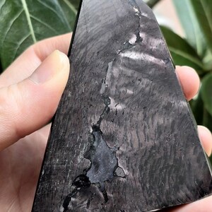 May include: A dark grey, triangular crystal with a textured surface and a unique pattern. The crystal has a crack running through the centre, adding to its natural beauty. The crystal is held in a hand.