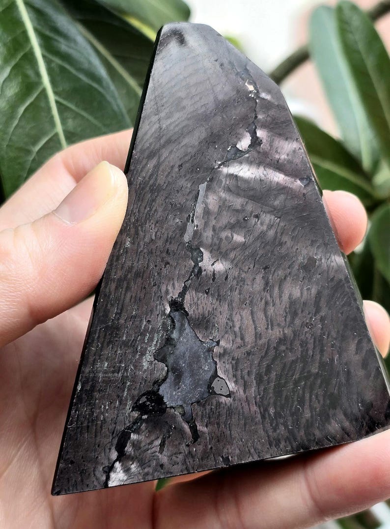May include: A dark grey, triangular crystal with a textured surface and a unique pattern. The crystal has a crack running through the centre, adding to its natural beauty. The crystal is held in a hand.