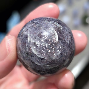 May include: A smooth, round, purple and silver sunstone sphere. The sphere has a shimmery, iridescent finish.
