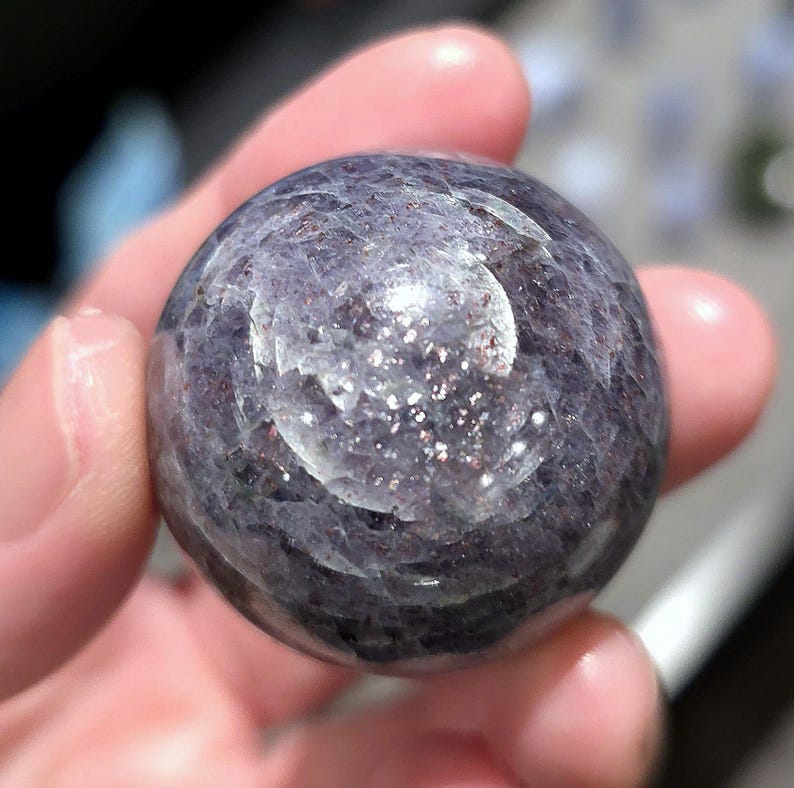 May include: A smooth, round, purple and silver sunstone sphere. The sphere has a shimmery, iridescent finish.