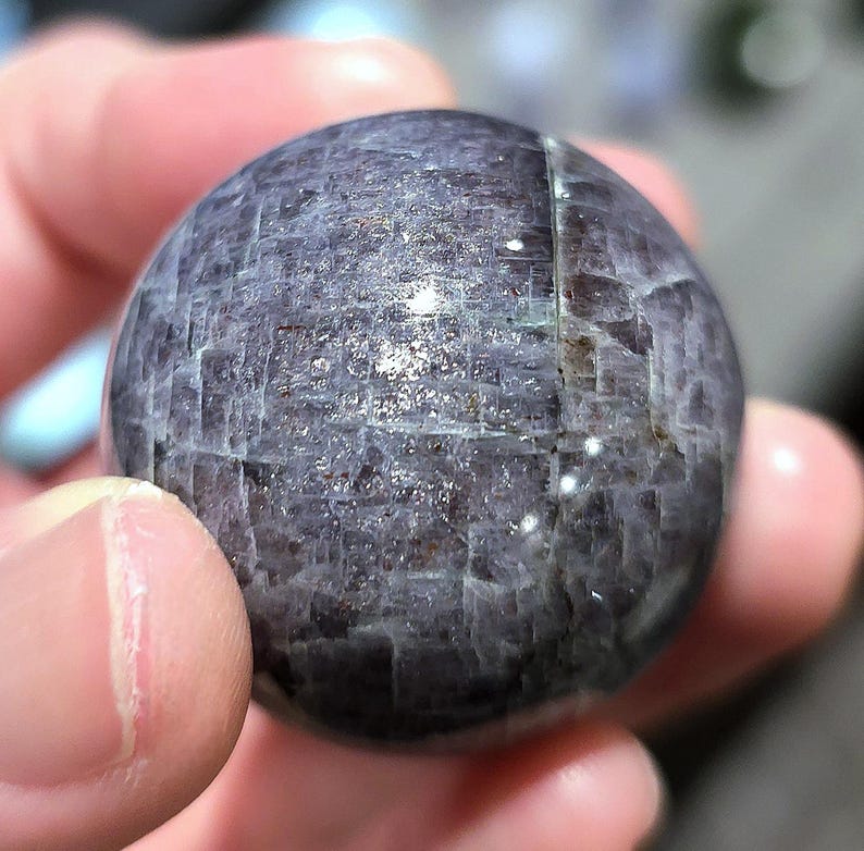 May include: A smooth, round, dark gray and silver sphere with a textured surface. The sphere is made of a natural material, possibly a crystal or gemstone.