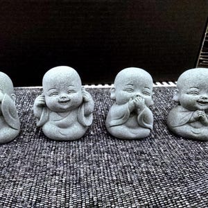 Op de afbeelding: Vier grijze stenen beeldjes van lachende, kale, mollige baby's. Elk beeldje staat in een andere pose, een bedekt zijn ogen, een bedekt zijn mond, een bedekt zijn oren en een houdt zijn handen voor zijn borst.