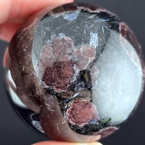 Puede incluir: Una gema esférica pulida, con una mezcla de secciones rojo oscuro y claras. La piedra tiene una superficie lisa y reflectante con patrones internos. La gema se sostiene entre los dedos sobre un fondo oscuro.