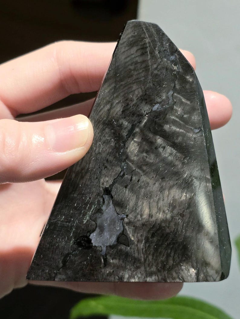 May include: A dark, triangular crystal with a polished surface. The crystal is primarily black with subtle grey and white veining, and a small, irregular cavity. The shape is reminiscent of a pyramid, suitable for display or collection.