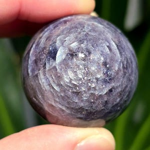 Puede incluir: Una esfera de color p&uacute;rpura y plateado con un patr&oacute;n de remolino. La esfera est&aacute; hecha de piedra natural y es lisa al tacto.