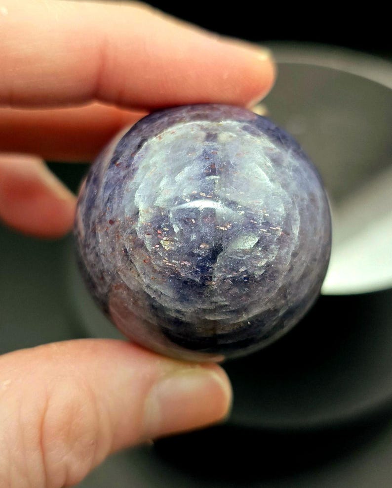 Puede incluir: Una esfera de piedra preciosa redonda, lisa, p&uacute;rpura y gris. La esfera tiene un patr&oacute;n de remolino y un acabado pulido brillante.