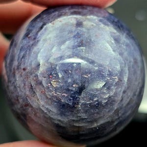 Puede incluir: Una piedra preciosa redonda, lisa, de color p&uacute;rpura y gris con un patr&oacute;n de remolino. La piedra preciosa est&aacute; siendo sostenida por una mano.