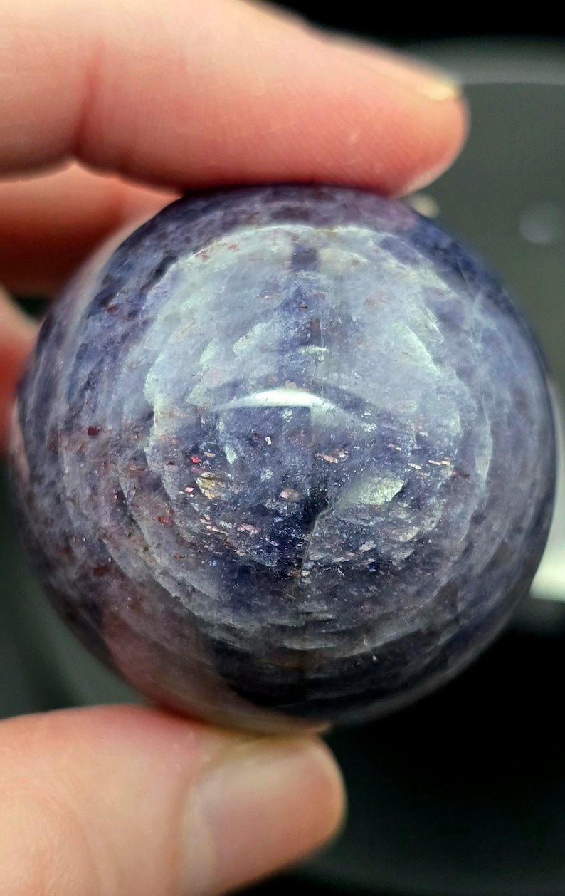 Puede incluir: Una piedra preciosa redonda, lisa, de color p&uacute;rpura y gris con un patr&oacute;n de remolino. La piedra preciosa est&aacute; siendo sostenida por una mano.