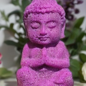 以下が含まれることがあります： 瞑想のポーズをとる鮮やかな紫色の仏像。像は質感のある多孔質の表面を持ち、木製の台座に置かれています。背景には緑の葉と紫の花があります。