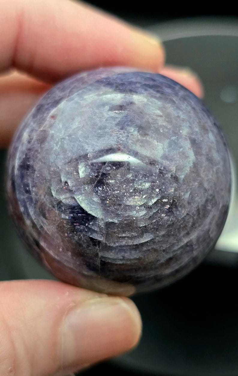 Puede incluir: Una esfera de piedra preciosa redonda y lisa, de color p&uacute;rpura y gris. La esfera tiene un patr&oacute;n de remolino de tonos claros y oscuros.