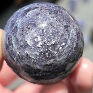 May include: A close-up of a round, purple and silver gemstone sphere. The sphere has a smooth, polished surface and a swirling pattern of silver flecks.