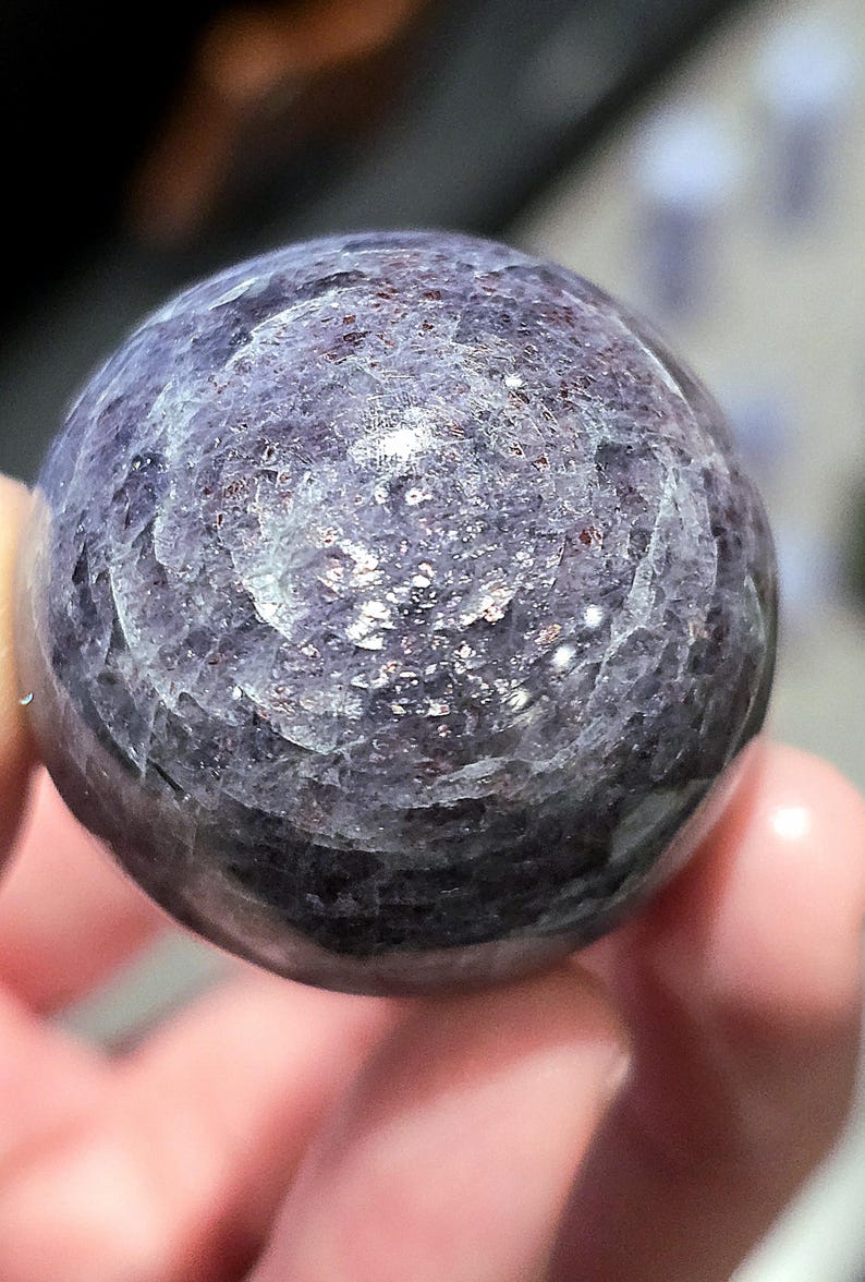 May include: A close-up of a round, purple and silver gemstone sphere. The sphere has a smooth, polished surface and a swirling pattern of silver flecks.