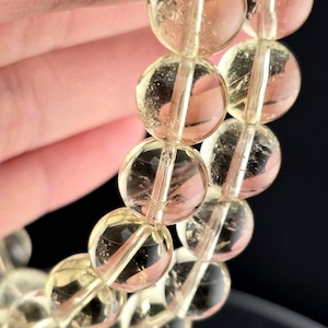 Op de afbeelding: Close-up van een armband gemaakt van doorschijnende, ronde, geel getinte kralen. De kralen zijn aan elkaar geregen en de armband wordt tegen een donkere achtergrond gehouden. De kralen hebben een gepolijste uitstraling.