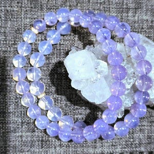 May include: Two beaded bracelets with light purple, round beads. The bracelets are next to a cluster of clear, crystalline quartz. The beads have a translucent quality, and the bracelets are arranged on a textured, gray surface.