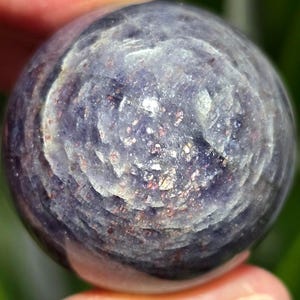 Puede incluir: Una esfera de piedra preciosa redonda, pulida, de color p&uacute;rpura y plateado. La esfera tiene un patr&oacute;n de remolino y un acabado brillante e iridiscente.