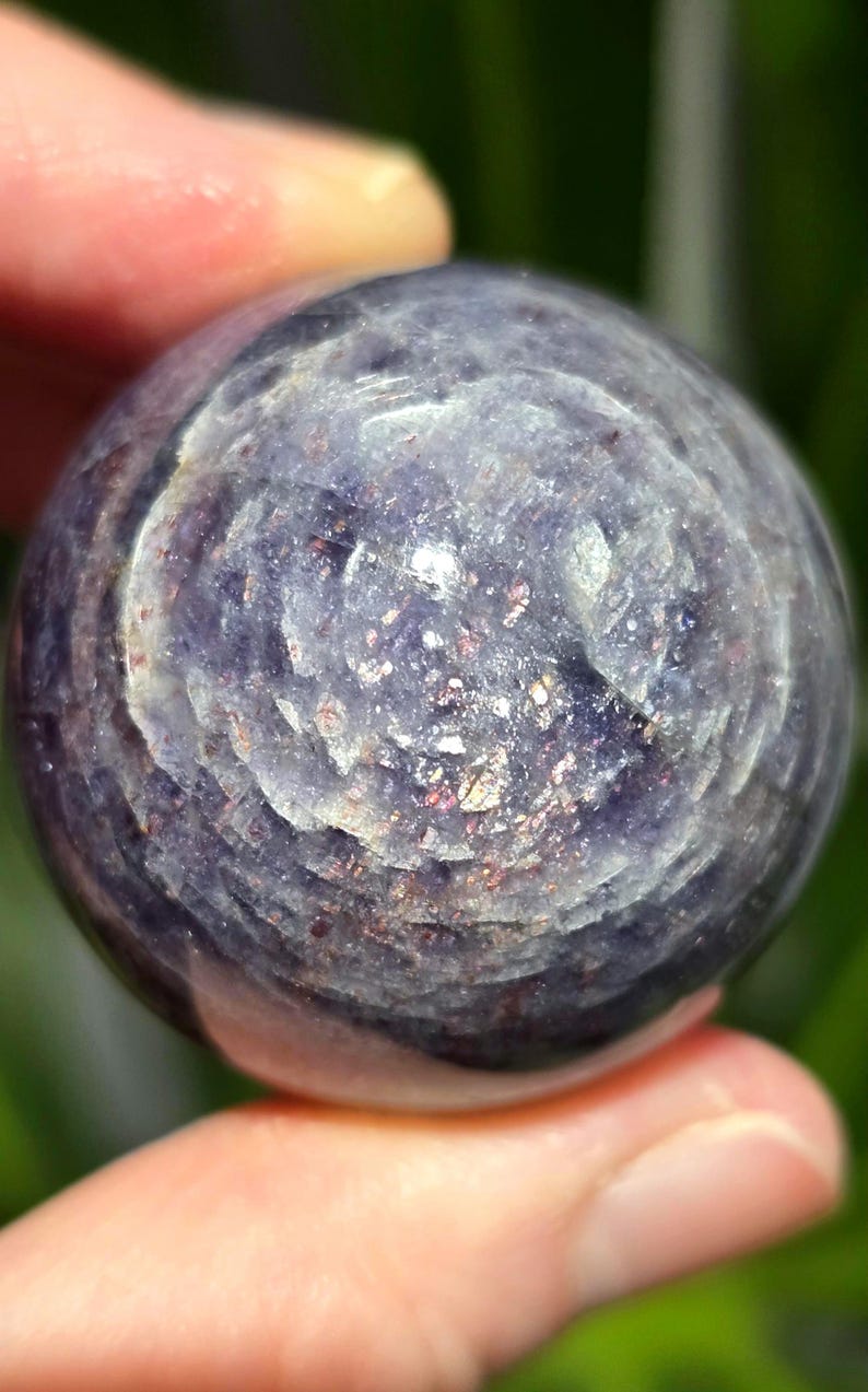 Puede incluir: Una esfera de piedra preciosa redonda, pulida, de color p&uacute;rpura y plateado. La esfera tiene un patr&oacute;n de remolino y un acabado brillante e iridiscente.