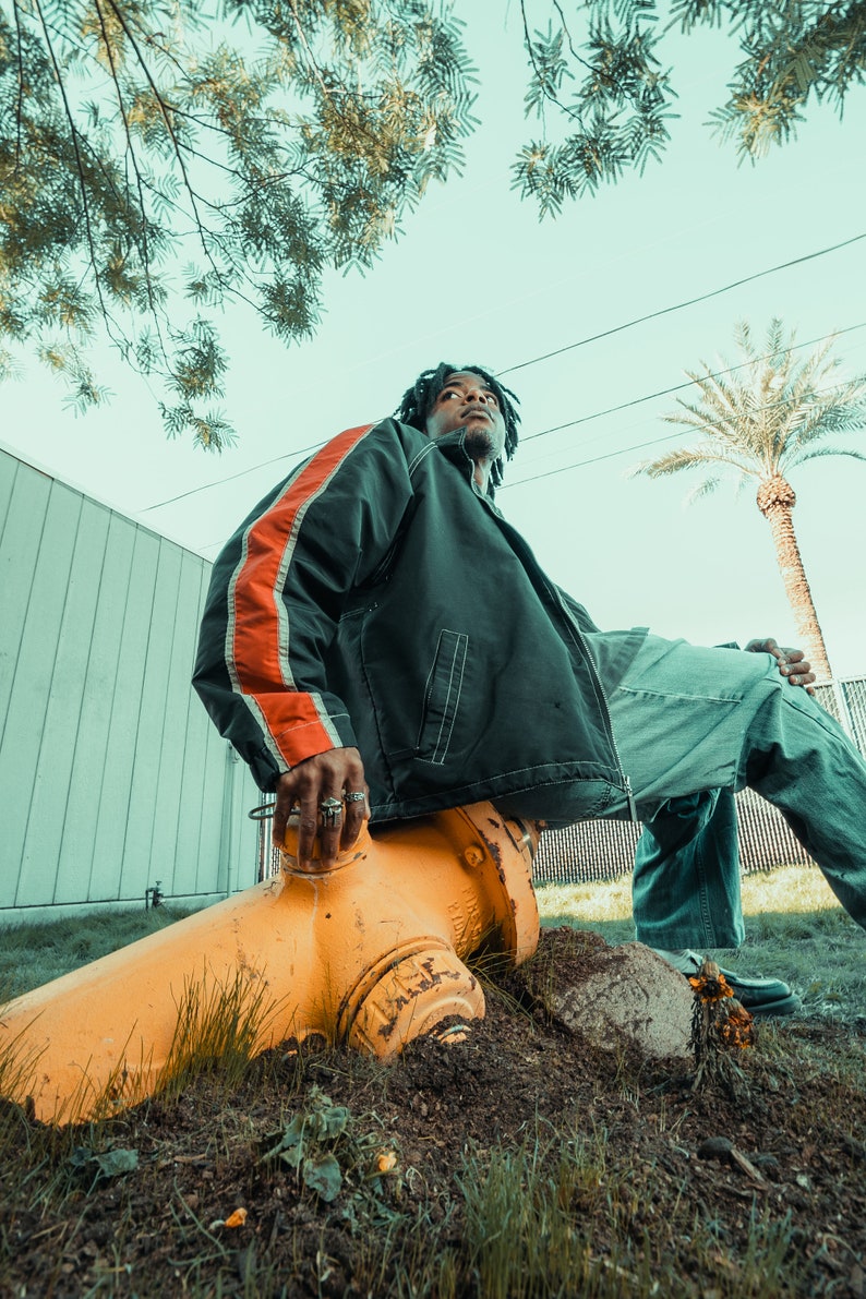 May include: A person wearing a black jacket with an orange stripe detail, and blue jeans, sitting on a yellow fire hydrant. The scene is outdoors with a palm tree and a clear sky in the background.