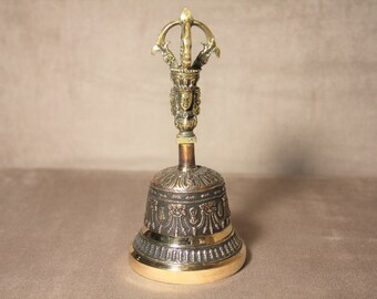 May include: A small, ornate brass bell with a detailed design. The bell has a handle with a stylized figure at the top.