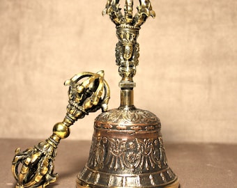 May include: A gold-colored bell with intricate carvings and a matching hand-held striker. The bell has a copper-colored interior and a crown-shaped top.