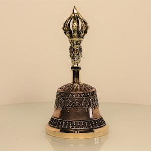 Könnte beinhalten: Eine traditionelle tibetische Glocke mit einem goldfarbenen Griff und filigranen Details. Die Glocke ist aus Kupfer und hat eine dunkelbraune Oberfläche.