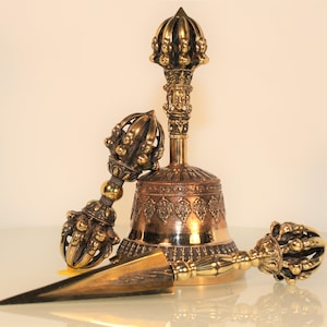 Könnte beinhalten: Eine Sammlung verzierter, bronzefarbener buddhistischer Ritualobjekte. Die Objekte umfassen eine Glocke, einen Vajra und einen Phurba. Die Glocke hat aufwendige Schnitzereien und einen detaillierten Griff. Vajra und Phurba sind ebenfalls sehr detailliert.
