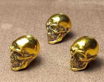 Op de afbeelding: Drie gouden schedelvormige kralen. De kralen zijn gedetailleerd met gezichtskenmerken en hebben een gladde, glanzende afwerking.