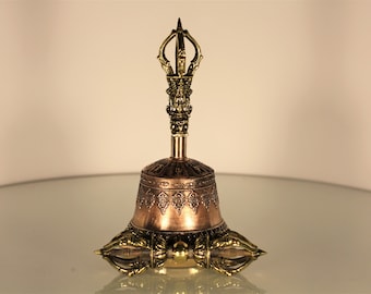 May include: A bronze-colored bell with intricate detailing. The bell features a decorative handle and a detailed base with a symmetrical design. The bell is likely made of metal and has a polished finish, suggesting it is a decorative or ceremonial item.