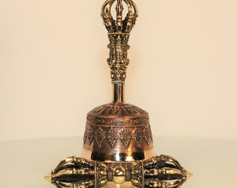 May include: A golden Tibetan bell with intricate carvings and a detailed handle featuring a face. The bell has a copper-colored base and a pair of ornate, hand-shaped handles.