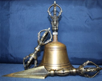 May include: A set of three antique brass Tibetan ritual objects. The set includes a bell, a vajra, and a dorje. The bell is decorated with a face and has a handle with a decorative top. The vajra and dorje are both decorated with intricate designs.