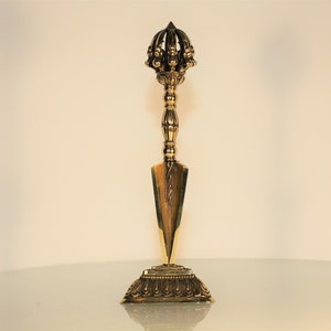 May include: A gold-colored, ornate ritual dagger with a detailed crown-like top and a triangular blade. The dagger is mounted on a decorative base, showcasing intricate patterns and designs. The object is likely used for ceremonial or spiritual purposes.
