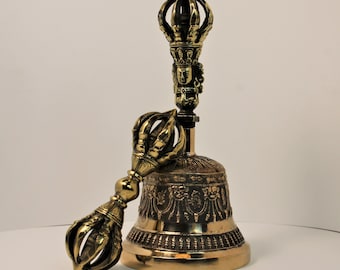 May include: A gold-colored Tibetan bell with a detailed design and a matching hand-held striker. The bell has a rounded shape with a decorative top and a handle. The striker has a curved handle and a decorative end.