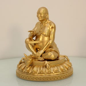 May include: A golden statue of a seated figure with a serene expression, pointing with one hand. The figure is adorned with a necklace and rests on a lotus-shaped base, with a dog-like creature at the base. The statue is a religious or spiritual item.