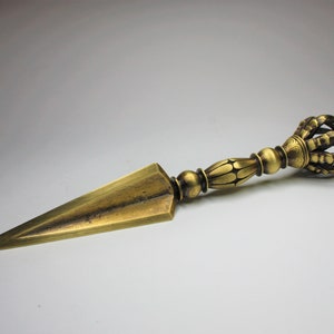 Op de afbeelding: Een gouden metalen dolk met een decoratief, versierd handvat. Het handvat is gevormd als een kroon met een puntige punt.