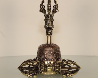 May include: A bronze-colored Tibetan bell and dorje set. The bell has intricate carvings and a handle with a detailed face. The dorje, or thunderbolt, is attached to the bell's base, featuring a symmetrical design with pointed ends.