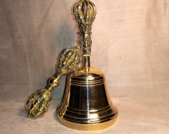 May include: A gold-colored bell with a decorative handle. The bell has a rounded shape and a wide opening. The handle is ornate and has a pointed top.