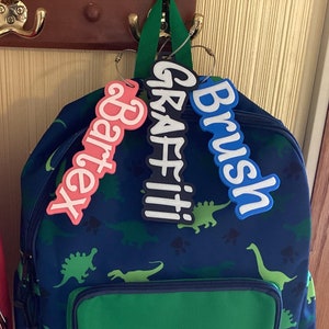 May include: Two backpacks hanging on hooks. The backpack on the left is pink with a green name tag that says "Script". The backpack on the right is blue with a green, pink, and blue name tags that say "Bartex", "Graffiti", and "Brush".