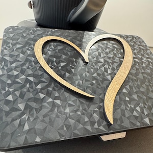 May include: A wooden heart-shaped decoration with a light brown finish rests on a dark gray surface with a geometric pattern. A black appliance with a silver handle is in the background.