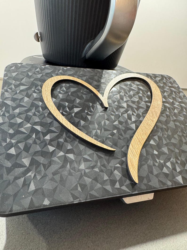 May include: A wooden heart-shaped decoration with a light brown finish rests on a dark gray surface with a geometric pattern. A black appliance with a silver handle is in the background.