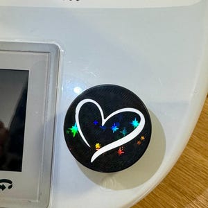 Könnte beinhalten: Ein schwarzer Handy-Griff mit einem weißen Herz-Design und schimmernden Sternen. Das Herz hat einen weißen Umriss und einen schwarzen Innenraum.