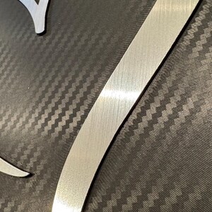 May include: Close-up of a metal art piece with a brushed silver finish. The design features a stylized letter "V" and a curved shape, set against a dark gray, textured background with a carbon fiber pattern.