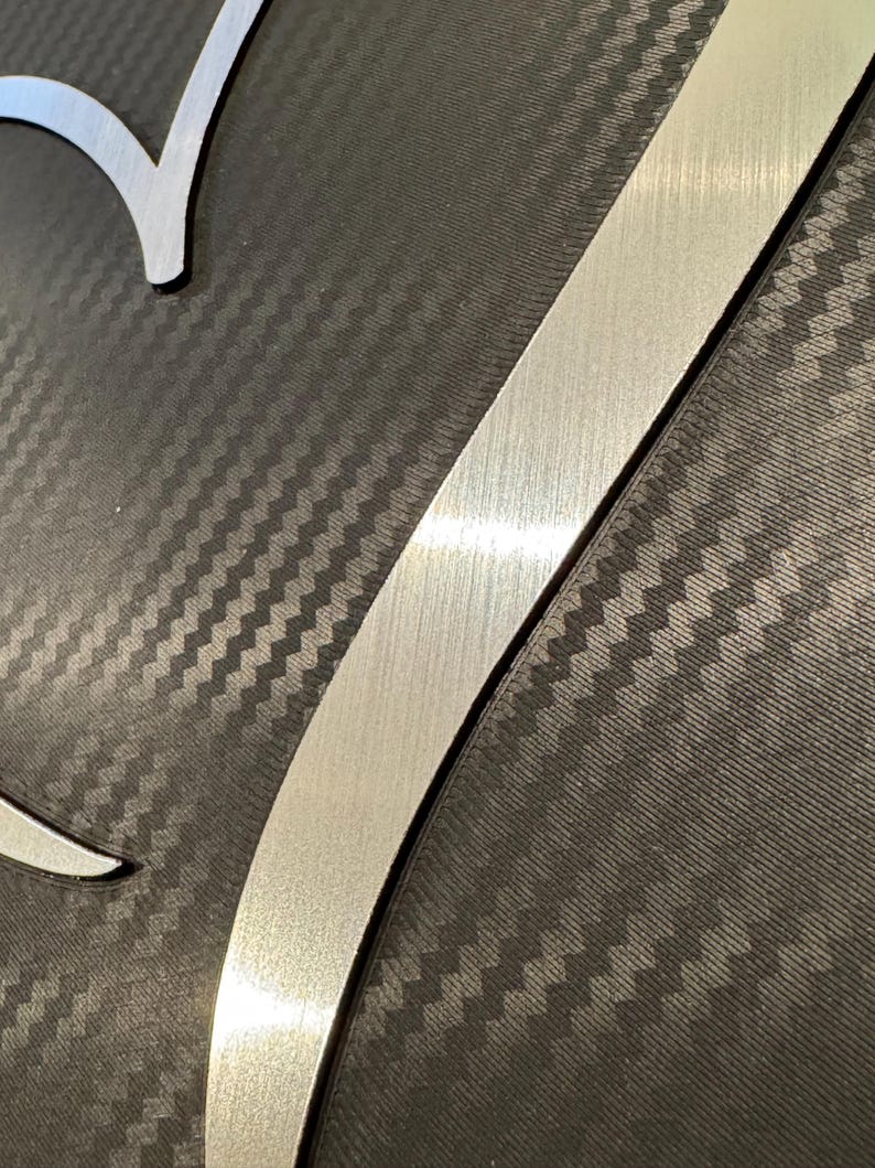May include: Close-up of a metal art piece with a brushed silver finish. The design features a stylized letter "V" and a curved shape, set against a dark gray, textured background with a carbon fiber pattern.