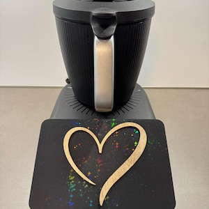 May include: A black electric kettle with a silver handle sits atop a base. A black rectangular surface with a wooden heart cutout and colorful star-like accents is in front of the kettle.