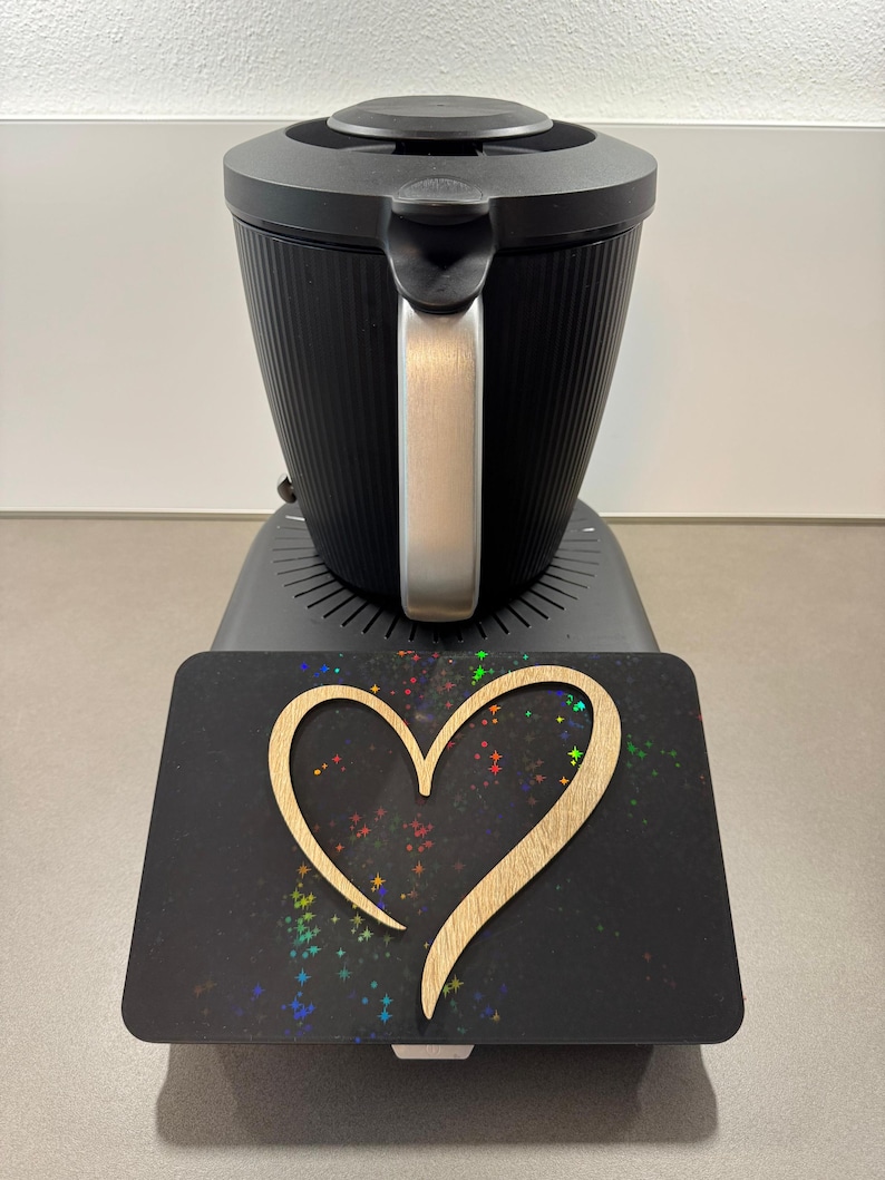 May include: A black electric kettle with a silver handle sits atop a base. A black rectangular surface with a wooden heart cutout and colorful star-like accents is in front of the kettle.