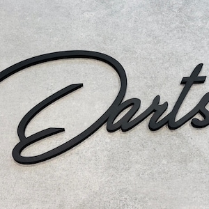 Könnte beinhalten: Ein schwarzes, dreidimensionales Wandschild mit dem Wort Darts in einer geschwungenen Schreibschrift. Die Buchstaben haben eine glatte, matte Oberfläche und sind in einem Winkel angeordnet, was eine klare und gut lesbare Wanddekoration ergibt.