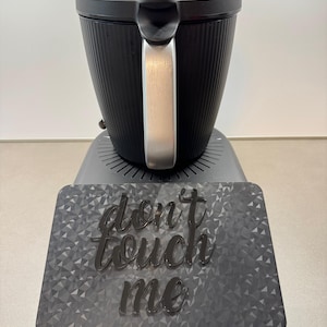 May include: A black appliance with a silver handle and a textured, ribbed design. The appliance sits on a gray base with a geometric pattern and the words "don't touch me" in a cursive font.