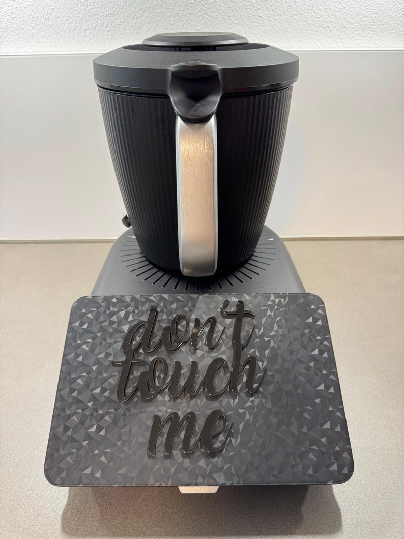 May include: A black appliance with a silver handle and a textured, ribbed design. The appliance sits on a gray base with a geometric pattern and the words "don't touch me" in a cursive font.