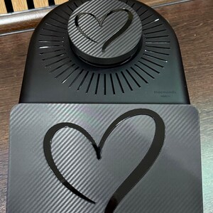 以下が含まれることがあります： カーボンファイバーのハートデザインが施された黒いキッチン家電。ハート型のアクセントが付いた丸い上部と、お揃いのハートが付いた長方形のベースがあります。「Thermomix」のブランド名がベースに表示されています。